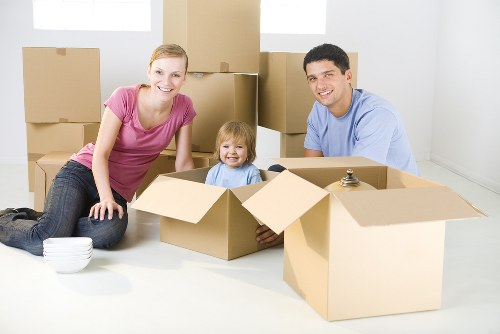 A family of three, including a woman, a man, and a young girl, sitting on a light-colored floor inside a residential space in Finchley after moving, surrounded by open cardboard boxes. The woman, dressed in a pink top and jeans, is smiling at the camera and leaning forward with her hands on the floor, while the young girl, with blonde hair and wearing a blue shirt, is sitting inside an open box, also smiling. The man, wearing a light blue shirt, is seated behind the girl, partly inside another box and smiling at the camera. In the background, there are several stacked cardboard boxes of various sizes, some closed and some open, indicating a recent move. The space has pale walls and natural lighting, suggesting a bright, clean house environment typical of a London suburb. The scene subtly reflects house relocation activities, associated with home removals services like those offered by Finchley Man with Van, in the nearby Finchley area postcode. The overall setting conveys a moment of unpacking and settling into a new home, with focus on household furniture and packing materials present in the room.