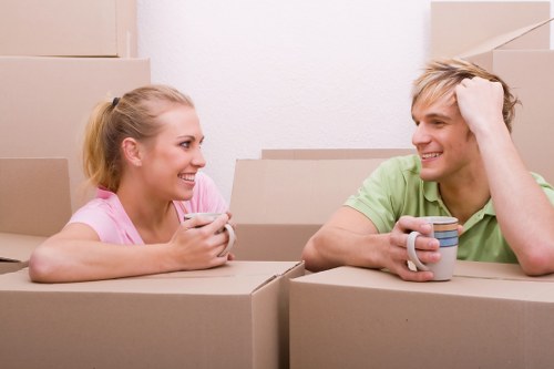 A young man and woman with light skin and blonde hair are sitting inside a room filled with cardboard moving boxes, which are stacked in the background and on either side of them. The woman, wearing a pink T-shirt, is smiling and holding a white mug, while the man, dressed in a green T-shirt, is also smiling and resting his head on his hand, holding a multicoloured mug. The environment appears to be a residential interior, possibly a hallway or living room in a house or flat, with plain white walls. The scene captures a moment of casual conversation or break during a house move, with visible packing materials indicating an ongoing relocation process. Light is evenly distributed, reflecting a well-lit indoor space typical of a house or flat in Finchley, London, area, evoking a friendly, relaxed atmosphere associated with professional removals services by Finchley Man with Van. The focus is on the interaction between the two individuals, with the packed boxes suggesting they are in the midst of a house or flat move or preparation for one.