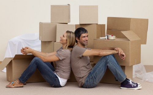 A man and a woman are seated back-to-back on a beige carpeted floor in a room, surrounded by stacked cardboard moving boxes of various sizes. The woman, positioned on the left, has blonde hair and is wearing a short-sleeved taupe top, blue jeans, and open-toed sandals, with her arms extended forward. The man, on the right, has dark hair and is dressed in a similar taupe top, blue jeans, and dark sneakers, with his arms extended to the side, appearing relaxed. The boxes behind them are plain brown cardboard, some open and others sealed, likely prepared for a house move. In the background, a white sheet covers an unknown object, and there are a few rolls of packing tape visible on top of the boxes. The scene suggests a residential environment, possibly a hallway or living room, during a house relocation, with natural or ambient lighting casting soft shadows. This setup aligns with the professional house removals services offered by Finchley Man with Van in London, serving areas including Finchley and nearby ZIP codes.