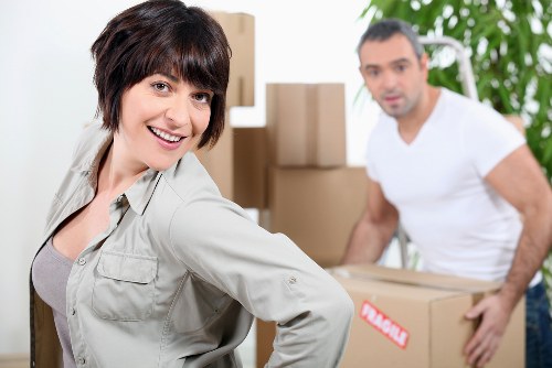 The image shows a woman with short dark hair, smiling and looking at the camera while wearing a light grey jacket, positioned in the foreground. Behind her, a man with short dark hair, wearing a plain white t-shirt, is slightly out of focus and lifting a medium-sized cardboard box. The background features several large stacked cardboard boxes, some with red and white labels, situated inside a residential space, likely a house or flat interior. To the right, there is a metal staircase railing and a leafy green plant, indicating a bright, well-lit environment typical of a home prepared for relocation. The scene captures a professional removals process, consistent with a house moving service operating in the Finchley area, as suggested by the context, with practical items such as boxes and modest furniture present, all with neutral tones and standard packing materials. Finchley Man with Van is associated with this arrangement, emphasizing local house removals expertise.