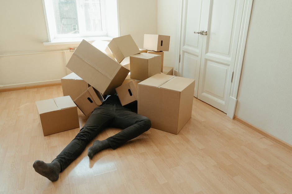 Inside a residential property during a home relocation process, a woman with long dark hair is seated on the floor, packing or unpacking a cardboard box. In the background, several stacked cardboard b
