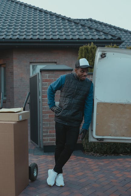 A man dressed in a blue jacket and dark trousers is seen loading a cardboard box into the back of a white panel van parked on a city street with a cobbled pavement. The van’s rear doors are open, re