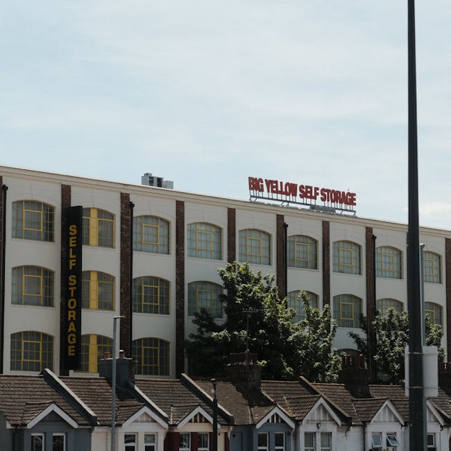 A mid-sized white moving van with a closed cargo area is parked on a narrow urban street, partially blocking the entrance to a building. The van is positioned close to the building's entrance, which i