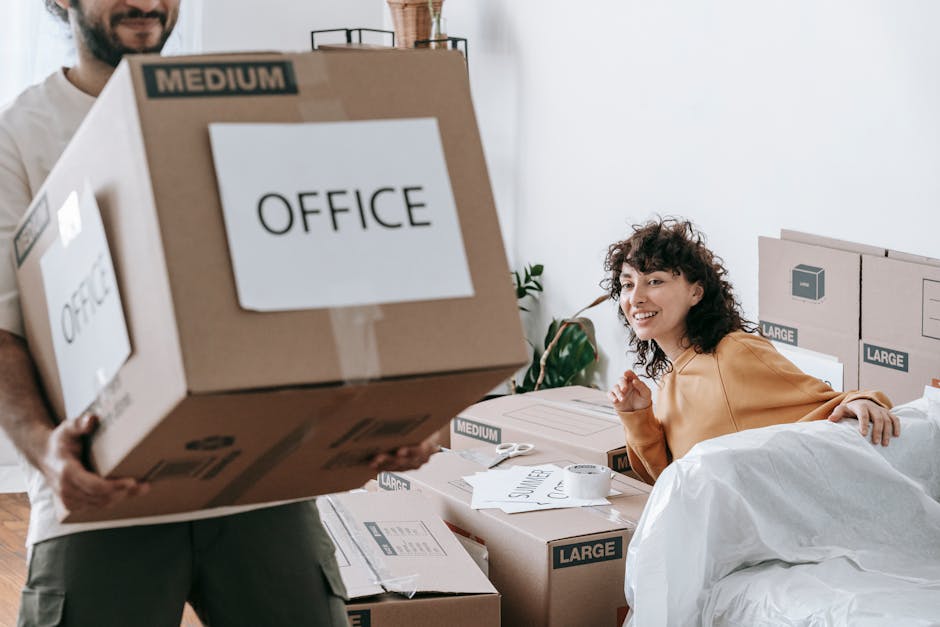 A man with curly black hair, glasses, and a beard is kneeling on the floor inside a well-lit room, engaged in packing or preparing furniture for a house removal. He is wearing a patterned short-sleeve